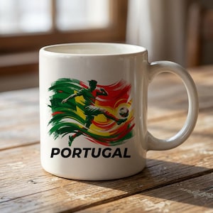 Puede incluir: Taza de cer&aacute;mica blanca con un jugador de f&uacute;tbol en acci&oacute;n, sobre un dise&ntilde;o verde, amarillo y rojo. La palabra "PORTUGAL" est&aacute; impresa en negro. La taza tiene un asa curva.