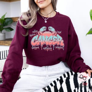 May include: A maroon sweatshirt featuring a colorful graphic design. The design includes the word "CANCER" in a graffiti-style font, along with a crab illustration and words like "caring" and "intuitive".