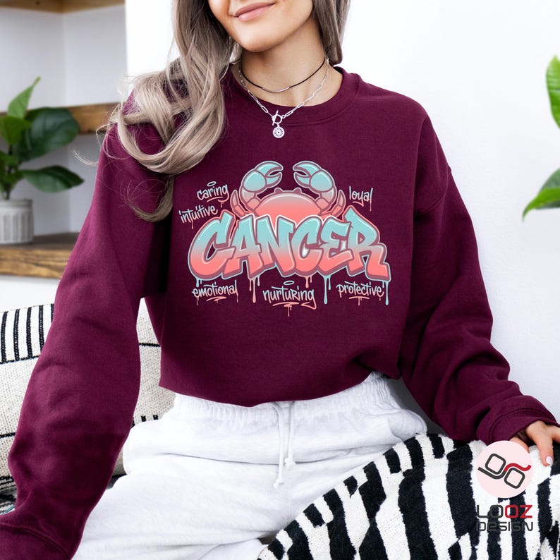 May include: A maroon sweatshirt featuring a colorful graphic design. The design includes the word "CANCER" in a graffiti-style font, along with a crab illustration and words like "caring" and "intuitive".