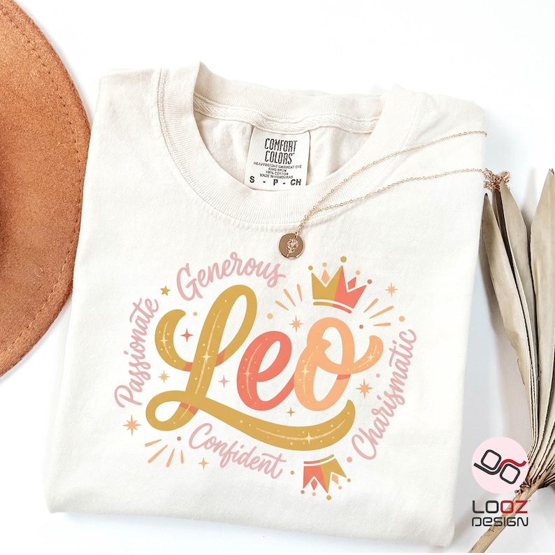 May include: Cream-colored t-shirt with a Leo zodiac design. The design features the word "Leo" in a stylized font, surrounded by words like "Passionate" and "Confident", with crown illustrations. A gold necklace with a pendant is draped over the shirt.