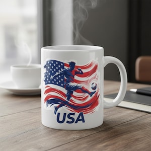 May include: White ceramic mug featuring a soccer player kicking a ball, set against a stylized American flag in red, white, and blue. The word "USA" is printed below the image. A cup of coffee sits in the background.