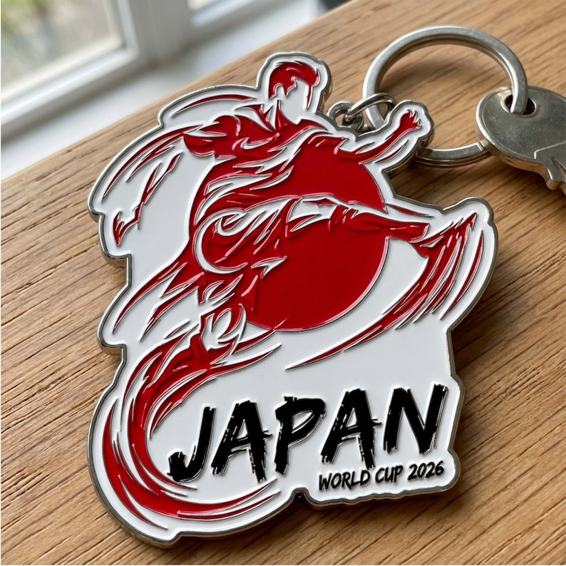 Może przedstawiać: Srebrny brelok do kluczy z czerwono-białą grafiką przedstawiającą postać w ruchu na tle czerwonego słońca. Słowo "JAPAN" jest wydrukowane na czarno, z napisem "WORLD CUP 2026" poniżej. Brelok jest przymocowany do k&oacute;łka na klucze.