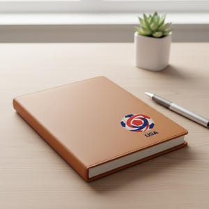 May include: A tan leather-bound notebook with a white-edged design. The notebook features a red, white, and blue logo with the word "USA" printed below it. A silver pen rests on the wooden surface.