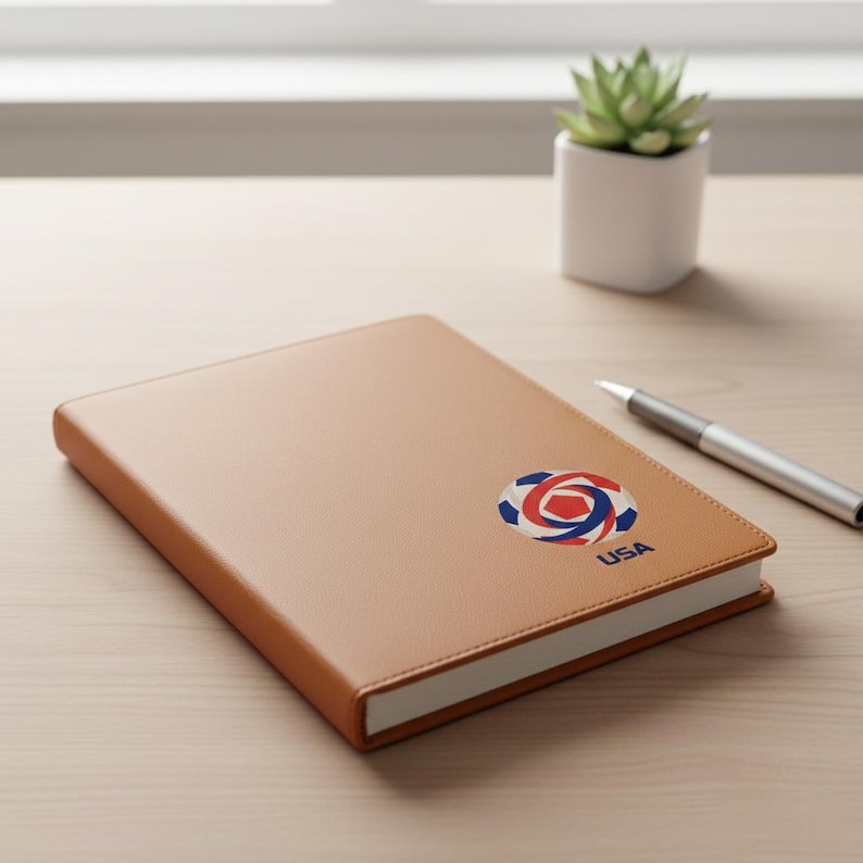May include: A tan leather-bound notebook with a white-edged design. The notebook features a red, white, and blue logo with the word "USA" printed below it. A silver pen rests on the wooden surface.