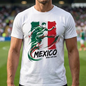 Może przedstawiać: Biała koszulka z graficznym przedstawieniem piłkarza w akcji, w barwach meksykańskiej flagi. Słowo "MEXICO" jest wyświetlane pogrubioną czcionką, a pod nim "WORLD CUP 2026".