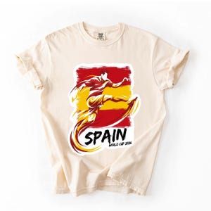 Może przedstawiać: Kremowa koszulka z grafiką piłkarza w akcji, w kolorach hiszpańskiej flagi, czerwonym i ż&oacute;łtym. Słowo "SPAIN" jest wydrukowane na czarno, z napisem "WORLD CUP 2026" poniżej.