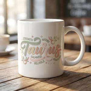 May include: White ceramic mug with a curved handle. The mug features the word "Taurus" in a stylized design with the words "Strength", "Calmness", "Reliable", "Nature", and "Grounded". The design is in pastel colors.