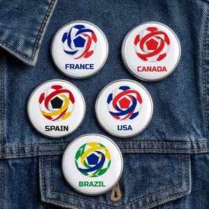 May include: Five round, white pinback buttons with colorful soccer ball designs and country names: France, Canada, Spain, USA, and Brazil. The buttons are on a blue denim jacket.