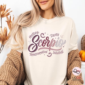 May include: Cream-colored t-shirt featuring the word "Scorpio" in a stylized script, with the words "Mystery," "Depth," "Transformation," and "Intuitive" surrounding it. The design incorporates purple and pink hues.
