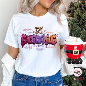 May include: White t-shirt with a colorful Sagittarius graphic. The design features the word "Sagittarius" in graffiti style, along with words like "adventurous" and "visionary". A Santa-themed mug is visible in the image.