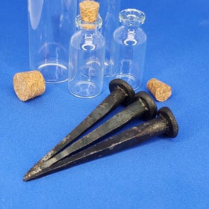 Puede incluir: Tres clavos oscuros de estilo antiguo con cabeza plana se muestran sobre una superficie azul, junto a pequeñas botellas de vidrio con tapones de corcho. Los clavos tienen un aspecto rústico y envejecido.