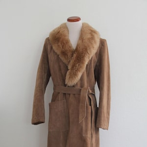 May include: A long, brown suede coat with a faux fur collar and a matching belt. The coat has two front pockets and is displayed on a wooden mannequin. The coat is a neutral color and appears to be vintage.
