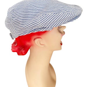 May include: A flat cap featuring a blue and white pinstripe pattern. The hat has a rounded crown and a short, stiff brim at the front. The fabric displays a subtle seersucker texture throughout, with a small adjustable strap visible at the back.