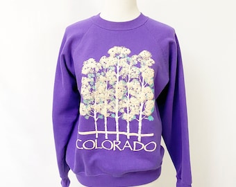 May include: A purple sweatshirt featuring a graphic of white trees with green and blue accents. The word "COLORADO" is printed in white below the trees. The long-sleeved top is displayed on a mannequin.