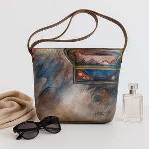 May include: A leather shoulder bag with a brown strap and a painted design in shades of blue, brown, and orange. The bag has a rectangular shape and a signature in the bottom right corner. Black sunglasses and a perfume bottle are next to the bag.