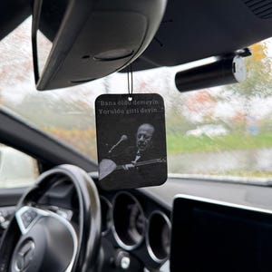 May include: Black rectangular car air freshener with a black string, hanging from a car's rearview mirror. The air freshener features a black and white image of a man playing a stringed instrument, with Turkish text.