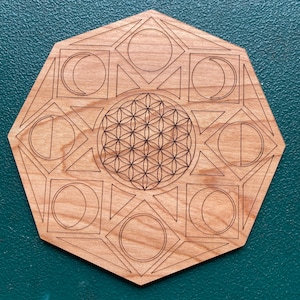 May include: A wooden, geometric design with etched lines. The design features a central flower of life pattern, surrounded by moons, circles, and geometric shapes. The wood has a natural, light brown color.
