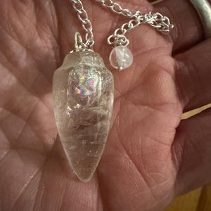 May include: A clear quartz crystal pendulum with a silver chain and a small, round, white stone. The quartz has a rainbow iridescence. The pendulum is used for divination and spiritual practices.