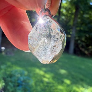 May include: A clear, raw, multifaceted quartz crystal pendant set in a metallic frame. The stone features internal fractures and natural inclusions that catch the sunlight, creating a bright lens flare at the top edge of the crystal structure.
