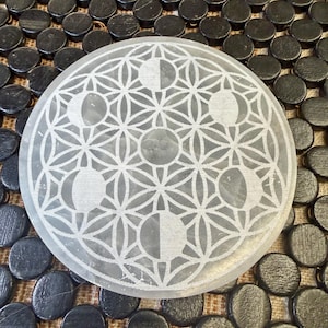 May include: A round, translucent selenite crystal plate with an etched design of interconnected circles and crescent moon shapes. The plate is set against a background of small, dark, circular objects.