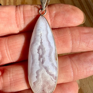 May include: A teardrop-shaped pendant featuring a pale lavender and white banded stone with a jagged, crystalline center. The stone is encased in a smooth silver-toned metal bezel setting with a triangular bail at the top for attachment.