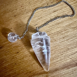May include: A clear, faceted, pointed crystal pendulum attached to a silver-toned metal chain. The chain features a small, round, clear crystal bead at the end. The crystal has visible internal inclusions and a smooth, polished surface texture.