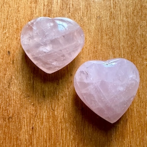 May include: Two heart-shaped rose quartz stones. The stones are a pale pink colour and have a translucent appearance. The hearts are smooth and polished, with visible natural veining. They rest on a wooden surface.