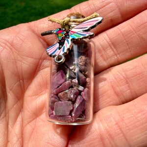 May include: A small clear glass bottle filled with raw, deep pink ruby gemstone chips. The bottle is sealed with a cork stopper and tied with a tan twine string. A metal hummingbird charm with colorful enamel wings in pink, blue, and yellow is attached.
