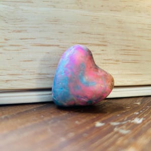 May include: A heart-shaped ornament with a swirling pattern of pink, blue, and purple hues. The object sits on a wooden surface, with a light-coloured wooden background.