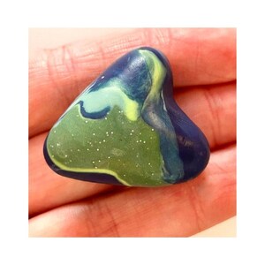 May include: A colorful, abstract stone with a rounded triangular shape. The stone features a blend of green, blue, and yellow hues, with a speckled, glittery texture on the green portion. The stone is held in a person's hand.