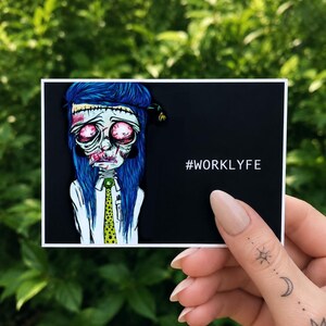 May include: A rectangular sticker with a black background featuring a stylized illustration of a zombie-like character with long blue hair, red eyes, and a white collared shirt with a yellow polka-dot tie. The text #WORKLYFE is printed in white.