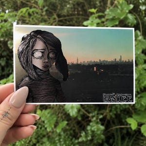 May include: A rectangular art print featuring a stylized illustration of a person with dark hair and large, pale eyes. The background depicts a city skyline at sunset with a gradient sky. The word HUSKEE is printed in a white graffiti-style font.