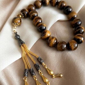 May include: A beaded prayer bracelet featuring round, polished tiger's eye stones. The bracelet has a decorative tassel with gold-colored chains and ornate metal accents. The beads are approximately 0.8 cm in diameter.