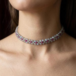 Könnte beinhalten: Nahaufnahme einer Halskette. Die Halskette besteht aus abwechselnden Reihen grauer und rosa Edelsteine. Die Edelsteine sind klein und rund und bilden ein zartes Muster. Die Halskette wird um den Hals getragen.
