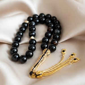 May include: Black prayer beads with gold-coloured detailing. The beads are spherical and polished, connected by gold-coloured spacers. The tassel and decorative elements are gold-coloured with black enamel accents. The background is a soft, neutral fabric.