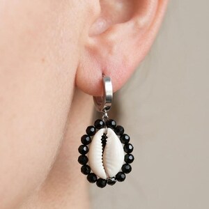 May include: A silver hoop earring with a pendant featuring a white seashell surrounded by a circle of black beads. The earring is shown worn on an ear.