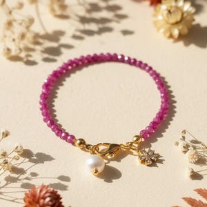 May include: A magenta beaded bracelet with a gold clasp, a white pearl, and a small, gold-coloured charm with clear crystals. The bracelet is displayed on a cream-coloured surface. The beads are approximately 0.3 cm in diameter.