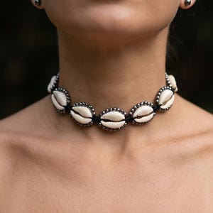 May include: A choker necklace featuring cream-colored seashells set in silver-toned metal. The shells are connected by a black cord, creating a striking contrast. The necklace is a piece of jewelry, perfect for adding a touch of coastal style.