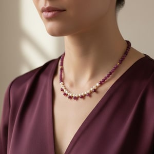 May include: A necklace featuring a strand of small, round, deep red beads alternating with white pearls. The necklace has a gold clasp and small gold accents. The woman is wearing a burgundy dress.