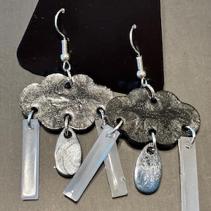 May include: A pair of dangle earrings featuring black glittery cloud-shaped charms. Each cloud is suspended from a silver-toned hook and hangs with three dangling elements: two translucent rectangular bars and one shimmering silver teardrop shape.