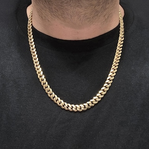 May include: A gold-colored curb chain necklace. The necklace is made of interlocking links and is worn around the neck. The background is a black shirt.