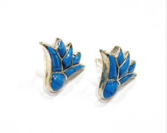 May include: A pair of silver-toned stud earrings. Each earring features a blue, flower-shaped design with a silver border. The earrings are set against a white background.