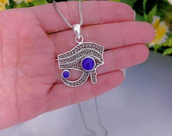 May include: A silver-toned pendant necklace featuring the Eye of Horus design. The pendant has intricate filigree detailing and two round, deep blue gemstones. The necklace chain is delicate and silver.