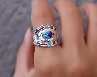 May include: Silver-toned ring with a colorful design. The ring features a central turquoise-colored stone, surrounded by red and blue accents. The band is adorned with additional red, blue, and turquoise details. The ring is worn on a finger.