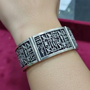 May include: Silver-toned cuff bracelet featuring engraved hieroglyphic symbols. The wide band bracelet is rectangular with a detailed pattern of ancient Egyptian writing. The jewelry is worn on the wrist and has a vintage or antique aesthetic.