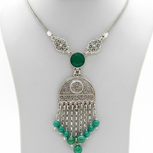 Può includere: Collana color argento con un ciondolo con una grande pietra verde e perline verdi più piccole. Il ciondolo ha un design ornato e dettagliato con un elemento circolare centrale e fili di perline pendenti. La collana è esposta su un manichino bianco.