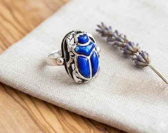 May include: A silver ring featuring a prominent beetle design with a vibrant, glossy blue enamel inlay on its back. The beetle is detailed with metallic silver legs and antennae, set on a polished silver band that encircles the finger.