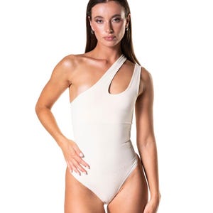 May include: A cream-colored one-shoulder bodysuit with a cutout detail on the neckline. The bodysuit has a high-cut leg and a form-fitting silhouette. The model is wearing the bodysuit.