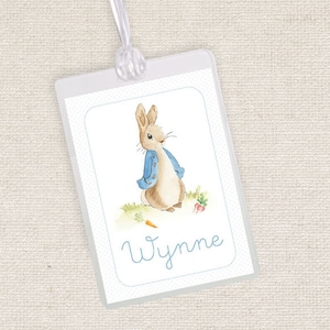 May include: A white luggage tag with a watercolor illustration of Peter Rabbit wearing a blue jacket. The tag has the name "Wynne" written in blue cursive.