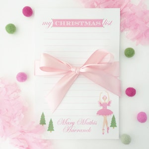 May include: A white notepad with the text "my CHRISTMAS list" in pink, a pink ribbon, and a ballerina illustration. The notepad is decorated with pink and green pom-poms and pink tissue paper.